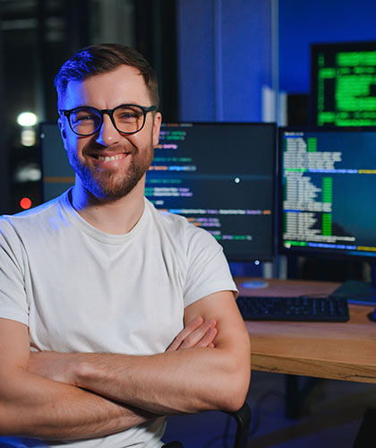 Hombre sonriente con gafas y brazos cruzados frente a pantallas con código